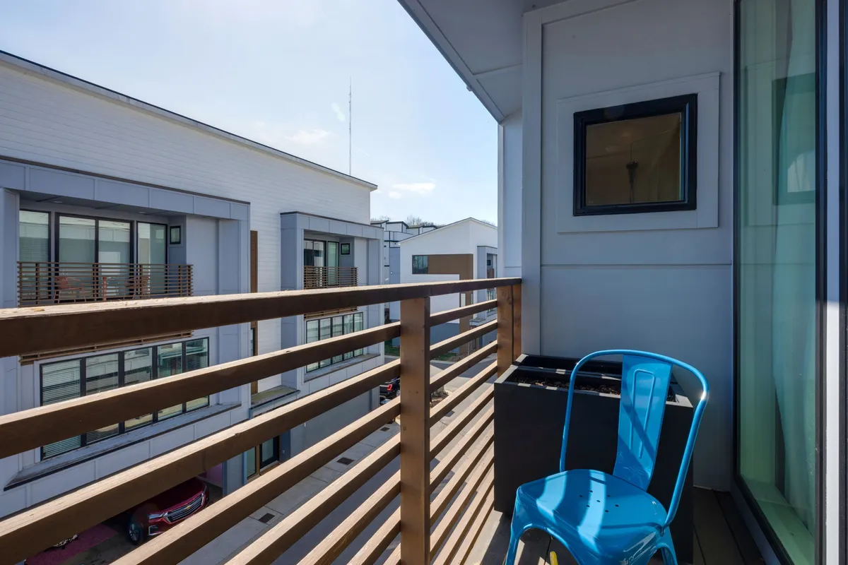 An exterior view of a private balcony with a blue chair and wooden railing, looking out over the neighborhood. The relaxed scene suggests a perfect spot to unwind after a day of exploring.