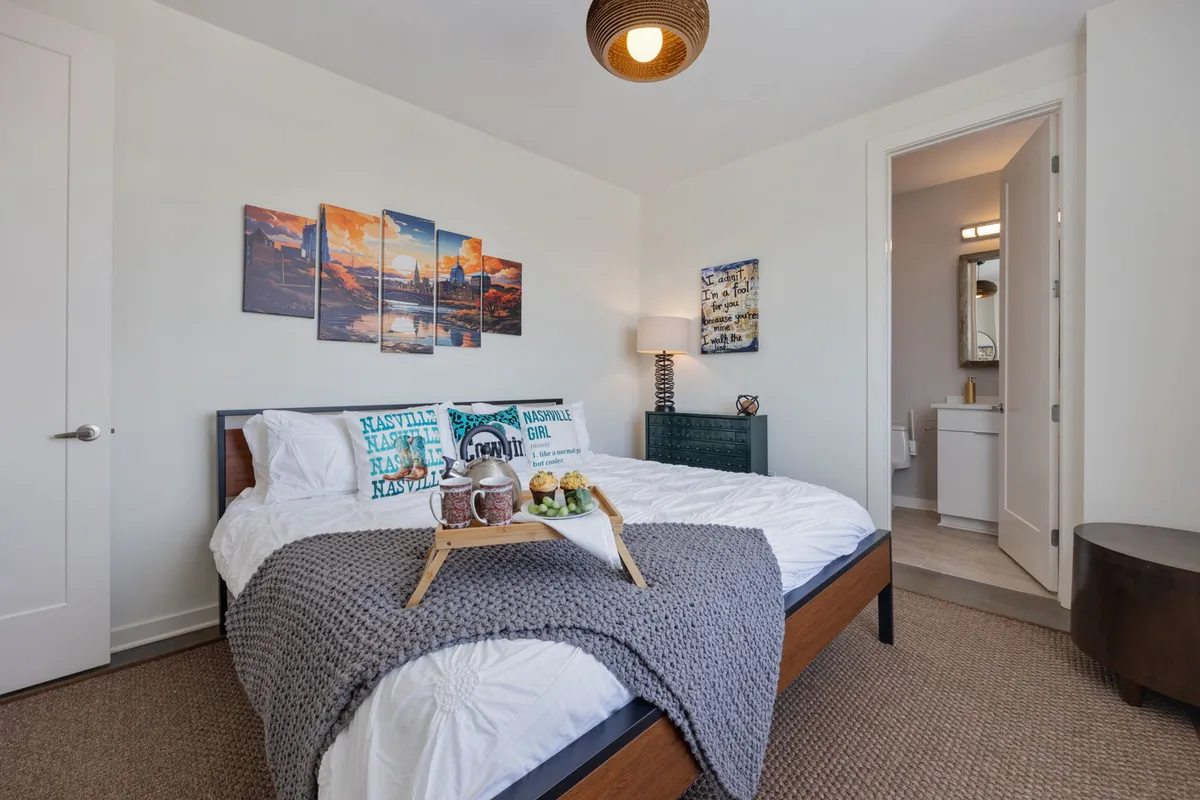 A stylish bedroom with a bed adorned with decorative pillows, artwork on the wall, a cozy nightstand with a lamp, and a tray set with fruit and drinks. The room exudes comfort and hospitality with soft lighting and artistic touches.