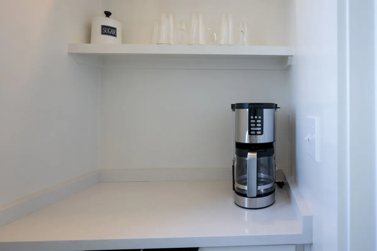A compact, practical beverage station featuring a coffee maker, sugar container, and glasses organized on shelf space. The setup is practical and thoughtfully arranged, suggesting the presence of standardized starter essentials for guests.