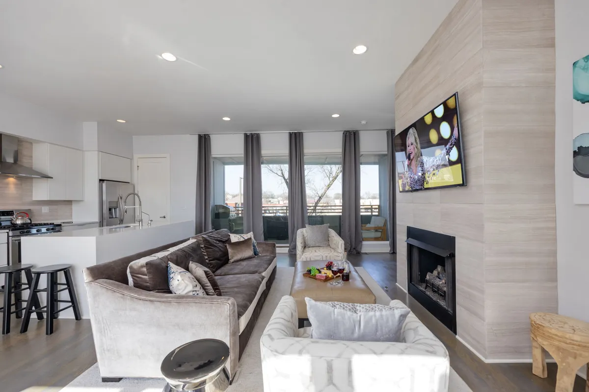 The image showcases a cozy, modern living space featuring a plush sectional sofa set around a fireplace and large TV. A nearby dining table invites gatherings, and the entire scene feels warm and inviting—perfect for relaxing, watching movies, or entertaining family and friends.