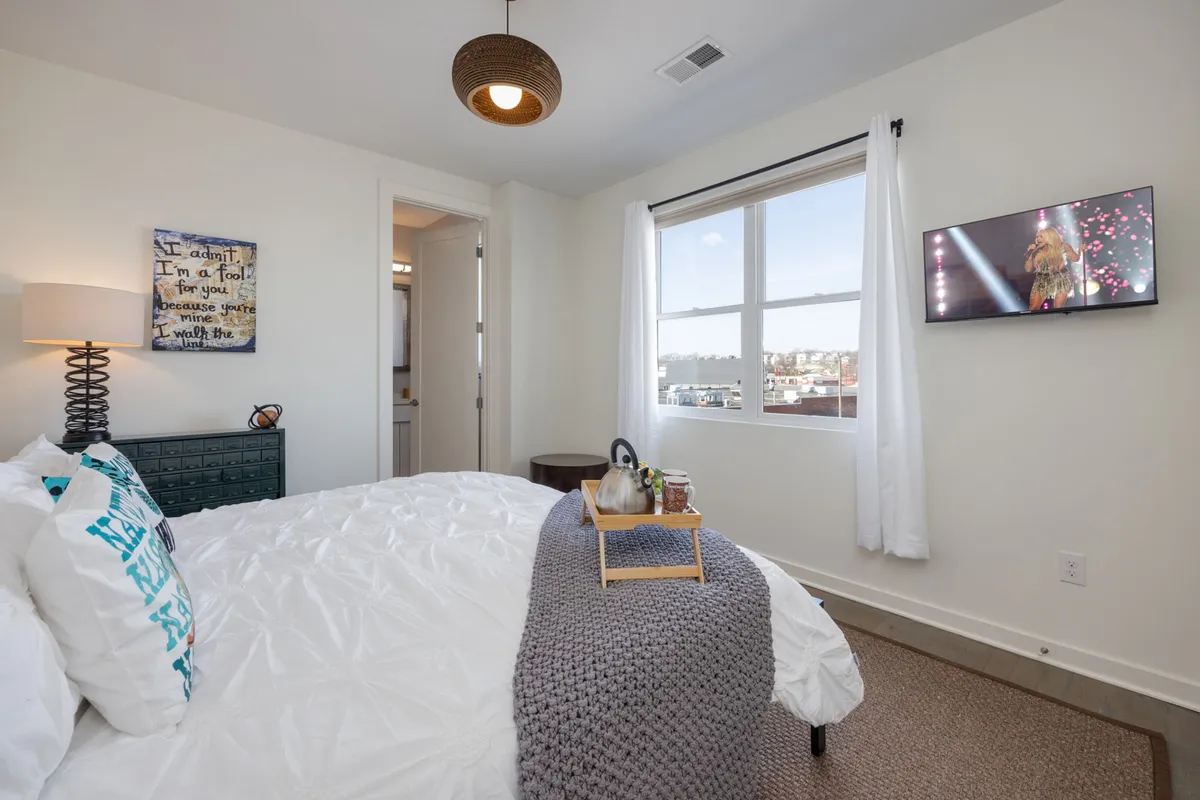 A bedroom with a cozy bed dressed in decorative pillows and a textured blanket, a lamp, wall art, and a mounted TV. The soft light and thoughtful layout create a casual, comfortable retreat.