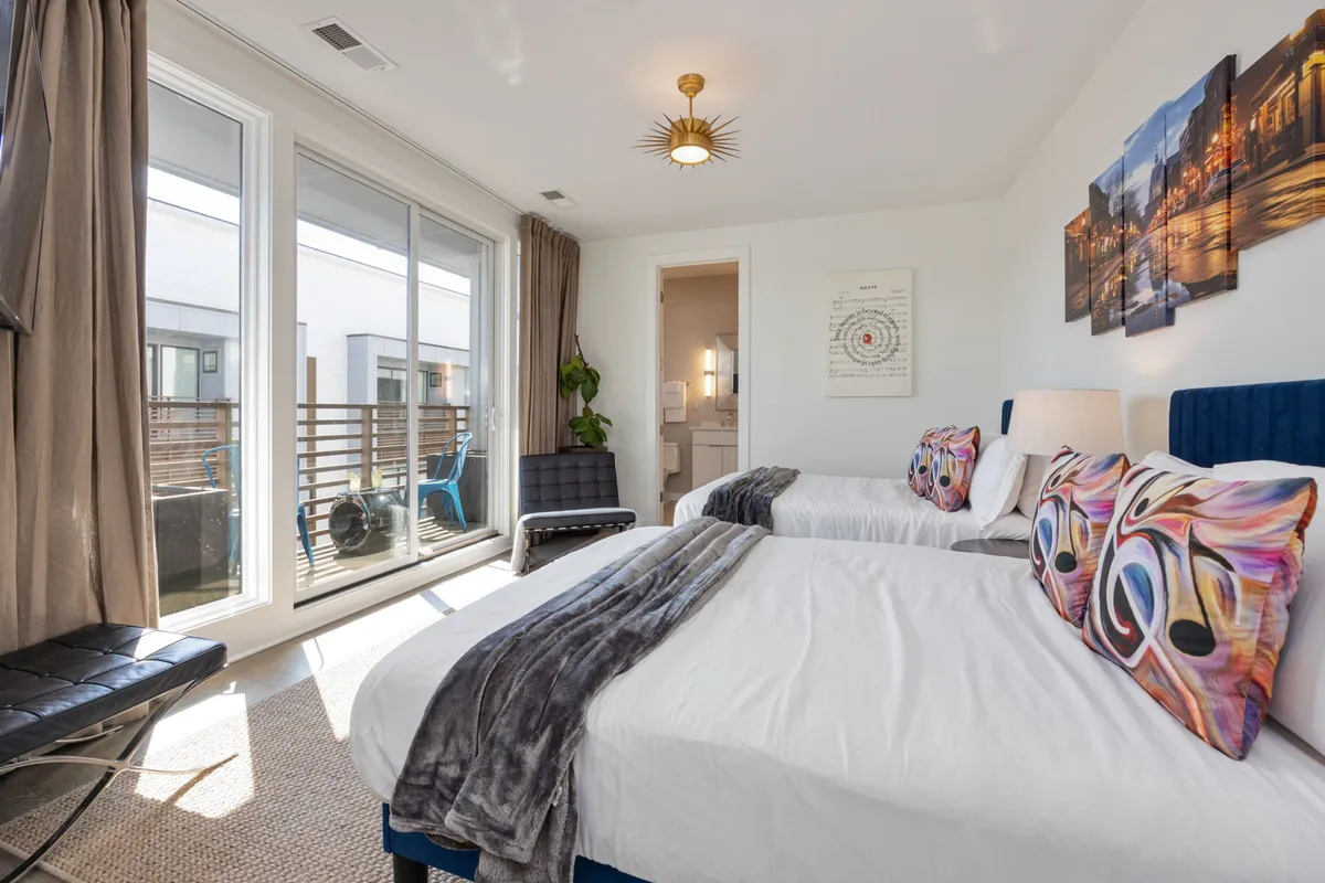 A cheerful bedroom featuring two full beds with colorful pillows, large windows letting in natural light, a stylish light fixture, and vibrant art on the wall. The room feels bright and ideal for friends or family to share.