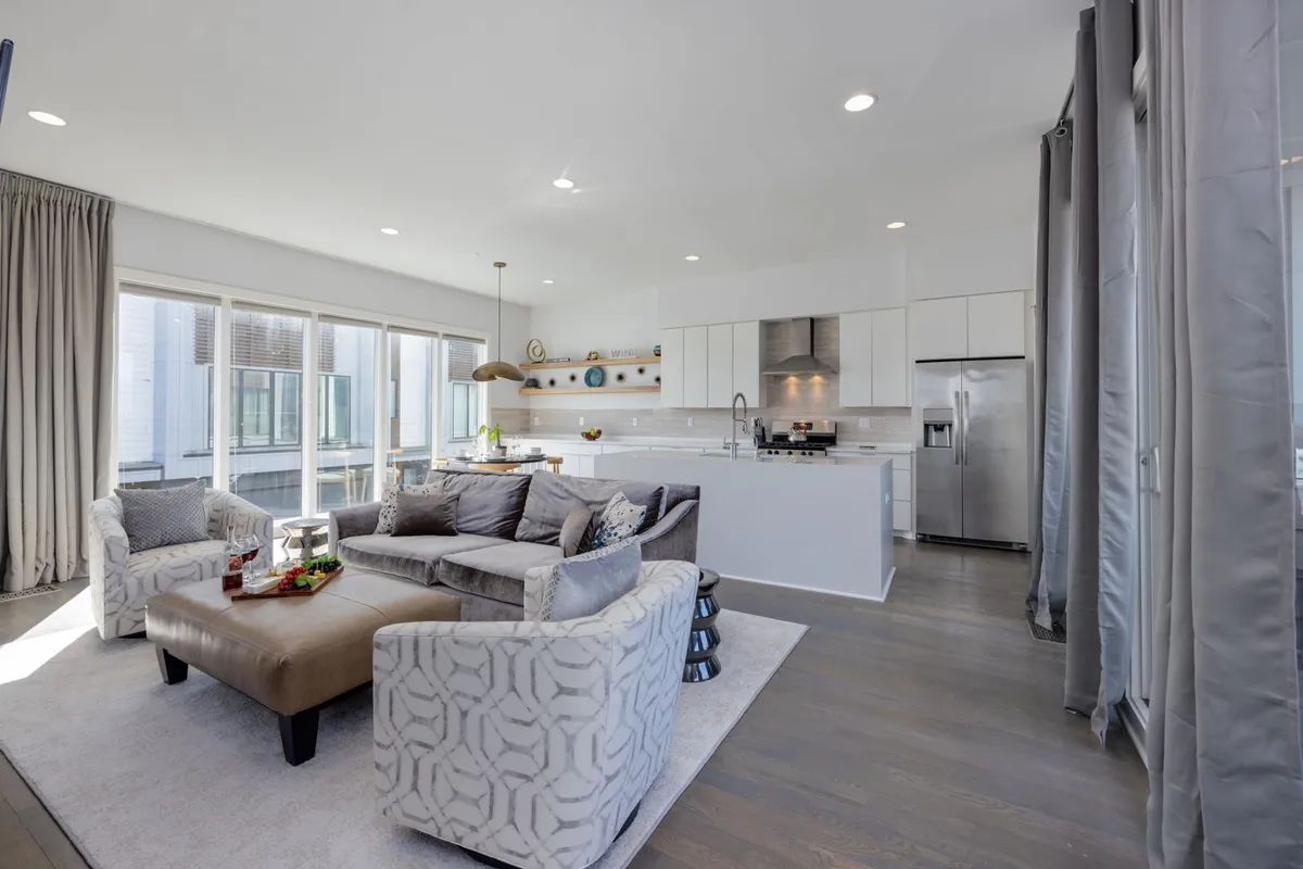 A welcoming, modern living space with a comfortable sofa, stylish accent chairs, a kitchen island, and large windows streaming in natural light. The room illustrates an inviting environment for relaxation, social gatherings, and everyday living.