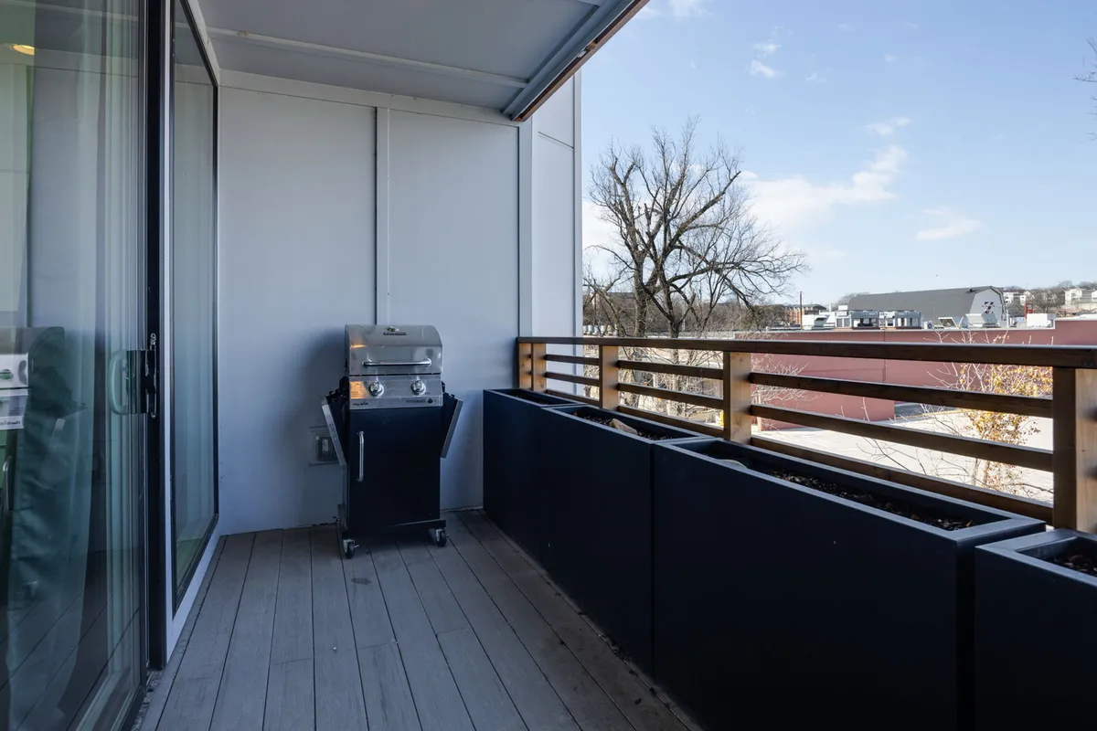 A private balcony scene with a barbecue grill, wooden railing, and planter boxes, suggesting a comfortable setting for outdoor dining and casual gatherings. The space is designed for both relaxation and entertaining, extending the living area outside.
