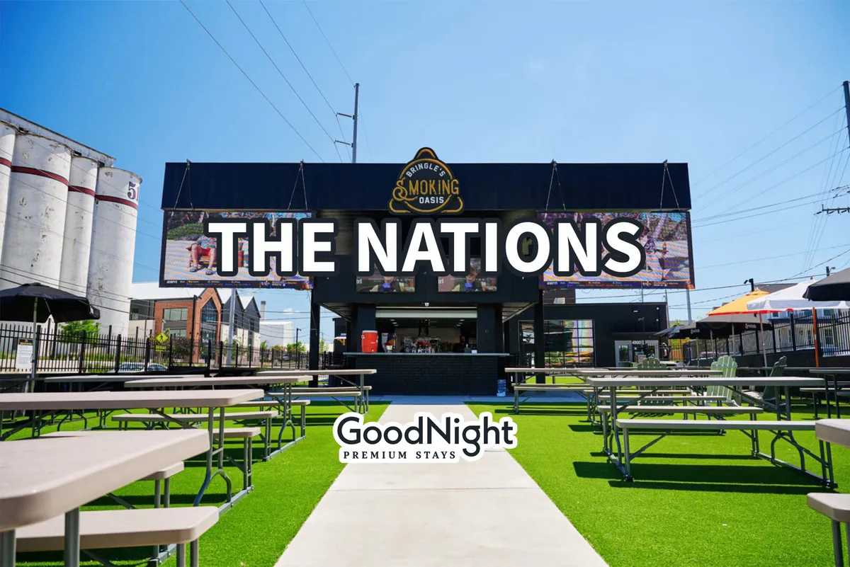 An outdoor scene featuring a large sign that reads 'THE NATIONS', with an outdoor seating area and a food service counter. The environment is lively, suggesting a vibrant gathering spot for locals and visitors alike.